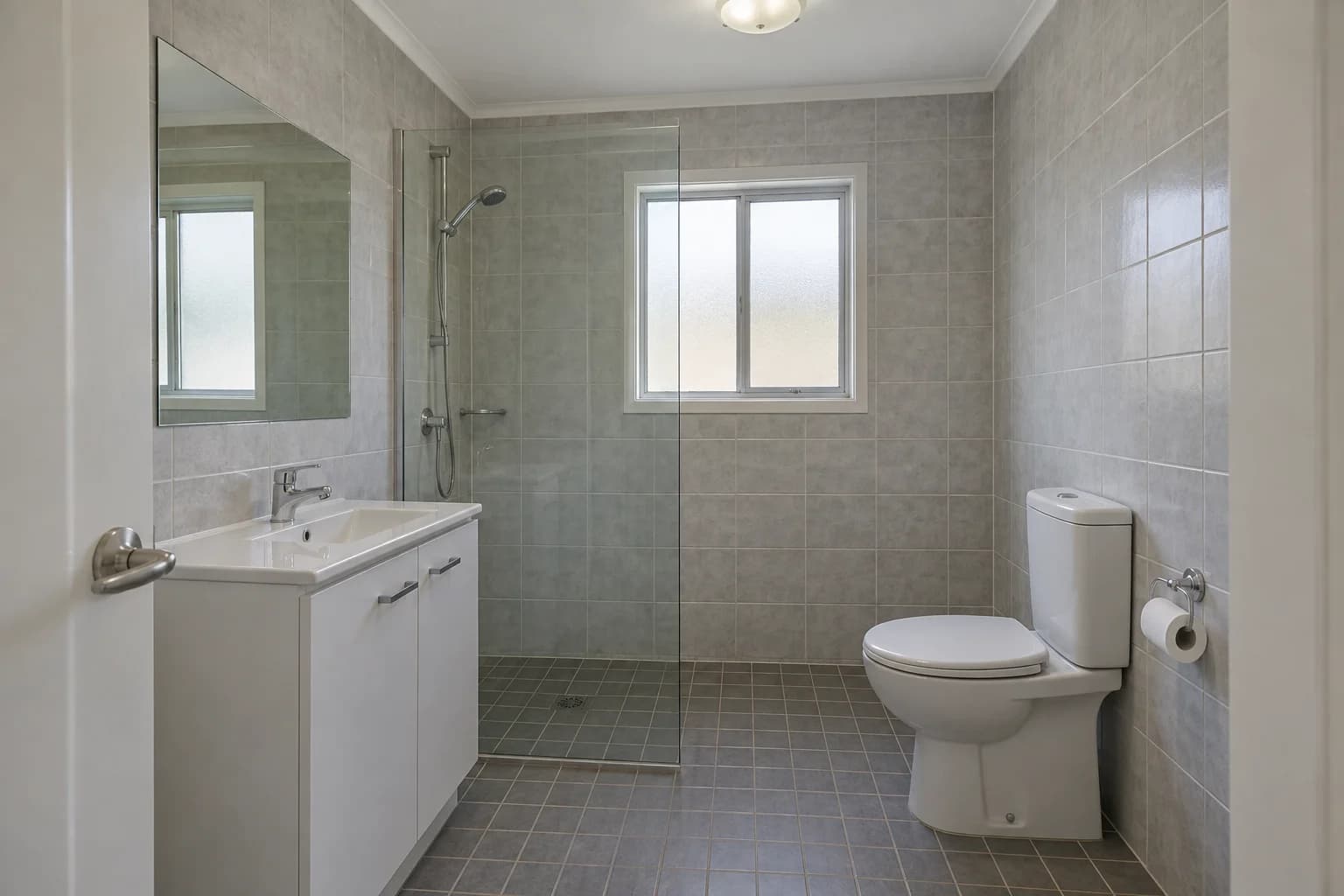 The starting bathroom with grey tiles, a white vanity, a rear shower and a toilet on the right.